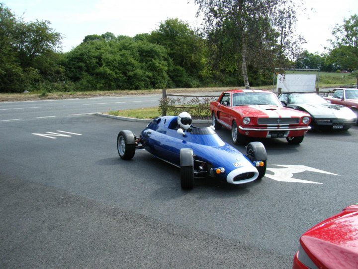 Breakfast Meeting, The Hare, Roxwell 4th June - Page 1 - Kent & Essex - PistonHeads - The image shows a small, blue race car parked in an open lot. The car is sleek, with a prominent nose cone and is equipped with slick tires. It is surrounded by a number of other vehicles, including cars and trucks that are painted in a variety of colors. The lot appears to be empty except for the racer, giving the car a central position in the frame. The sky is blue with some clouds, suggesting a clear day.