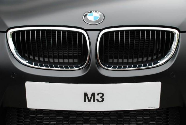 RE: PistonHeads Sunday Service: 25/07/10 - Page 9 - Events/Meetings/Travel - PistonHeads - The image features a close-up view of the front grille of a grey BMW car. The grille has a honeycomb pattern, typical of the BMW design. In the center of the grille, there is the BMW logo, which represents a quail with an arrow through it on a blue background. Below the grille, in front of the bumper, there's a clear white license plate with the letters "M3" printed on it. The style of the image is realistic and it focuses on the details of the car's front view.