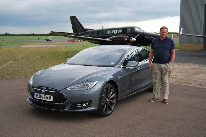 Friend's Tesla S  - Page 1 - EV and Alternative Fuels - PistonHeads - The image features two individuals standing outside, positioned next to each other with the man on the right standing slightly behind the other. Directly between them, on a brown cement surface, is a Tesla Model S sedan car. Above the cars, an ultralight aircraft equipped with a black and red helicopter is visible, parked in the background. The cloudy sky provides a bit of soft diffused light on the scene, and the overall setting appears to be an airfield.