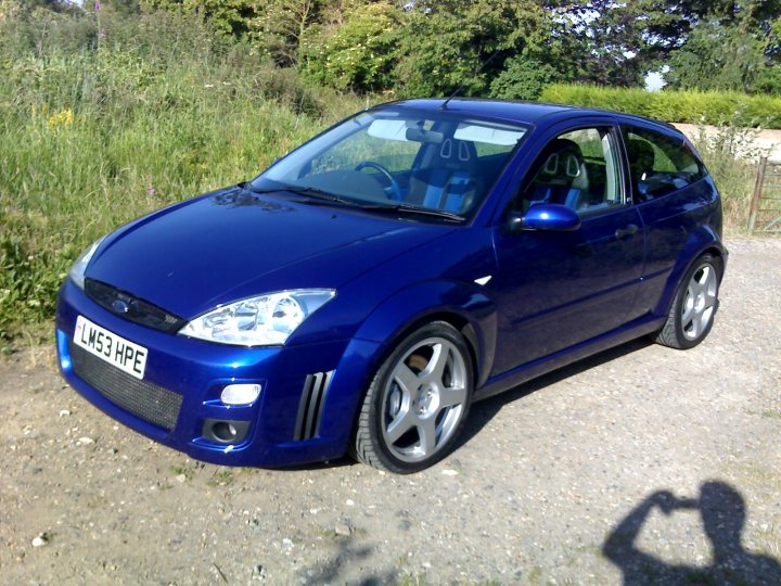 Pistonheads - The image shows a blue hatchback parked on a gravel driveway. The vehicle appears to be a compact or subcompact Fiat, evident by its distinctive front grill design. The wheels of the car have visible alloy rims, and the car itself is black, not blue. In the background, there is a dense foliage and a fence, suggesting a rural or semi-rural setting. On the right side of the image, there is a partial shadow of someone, likely the photographer. The overall scene appears to be a backdrop for car photography, characterized by a clear sky and a calm environment.