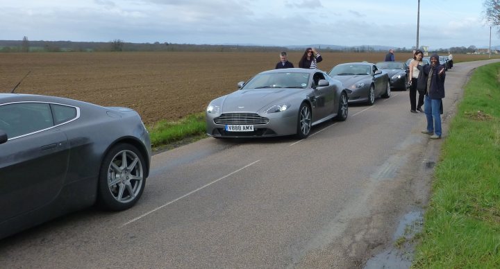 How about an Aston photo thread! - Page 68 - Aston Martin - PistonHeads - The image depicts a scenic road lined with green fields on both sides, where two elegant sports cars, a silver and a gray one, are driving. A handful of people are standing beside the cars, seemingly waiting to board. The passengers appear to be dressed for a journey or event, with ties being visible on at least two individuals. The overall atmosphere of the scene is that of an organized trip or excursion.