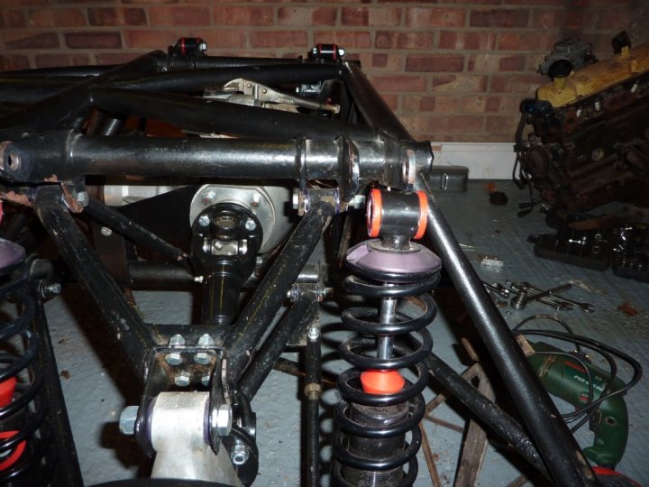 help please, with twin rear springs/shocks on S3 Vixen  - Page 1 - Classics - PistonHeads - The image shows a partly disassembled car. In the foreground, there's a metal object resembling a suspension piece, which appears to have a spring inside. The spring is sitting on a metal frame, possibly forming part of a shock absorber system. There are other mechanical components scattered on the ground, suggesting the vehicle has been worked on or taken apart. The background is slightly blurred but reveals a section of a red brick wall. The overall setting seems to be a garage or workshop environment. The image is of low resolution and has an overall monochromatic color scheme, emphasizing the metallic components.