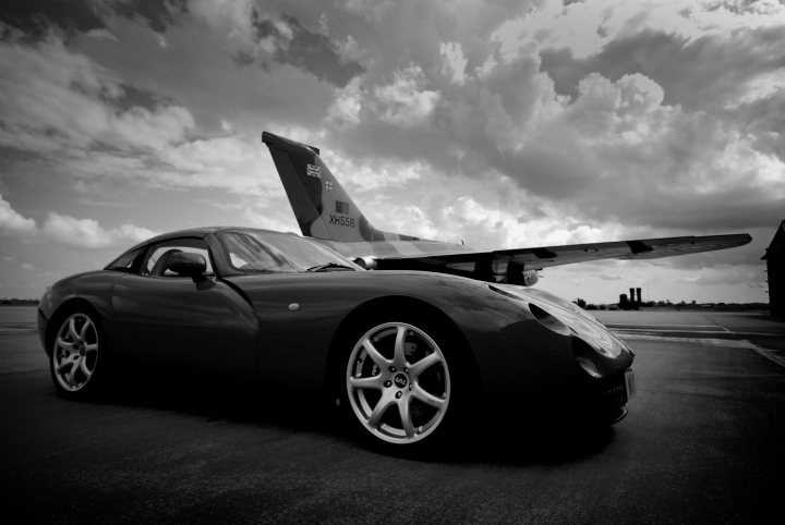 Your Best Tuscan Pic... - Page 8 - Tuscan - PistonHeads - The image captures a scene where a striking blue race car is parked on an airport tarmac. Its sleek design and low stance give it an imposing presence. The aircraft in the background, a large jet airliner, dwarfs the car by comparison. The gray, cloudy sky adds a dramatic backdrop to the scene. The composition of the image, with the races car in the foreground and the airplane behind, creates a sense of depth and scale. The lack of color only accentuates the contrast and composition of the image.