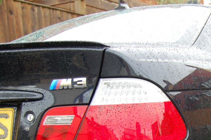 Lucky Luciano detailed my motor! - Page 1 - South West - PistonHeads - The image shows a close-up view of the back of a vehicle, featuring a BMW logo. The car is black with a red tail light, and is seen on a rainy day, with droplets on its surface indicating it has just driven through rain. The reflections on the wet surface create abstract patterns around the car, adding a dynamic element to the scene. The license plate of the vehicle is partially obscured by the droplets.