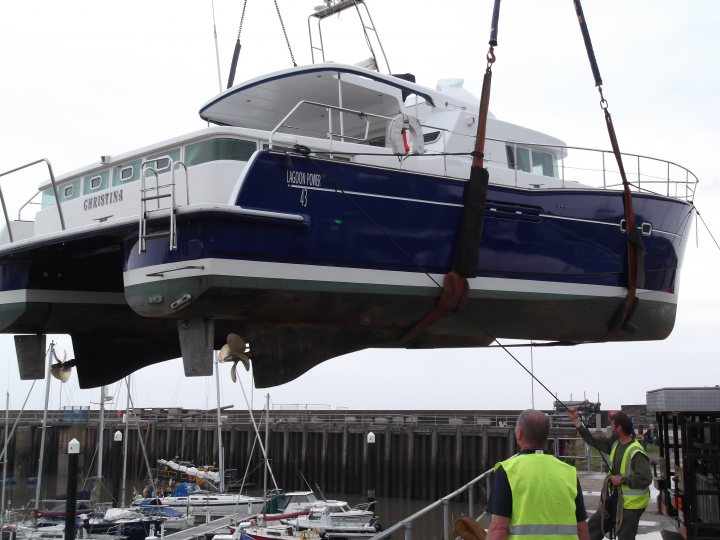 Wraps - Page 1 - Noble - PistonHeads - This image shows a large blue and white yacht being lifted for maintenance or storage by a crane. The yacht appears to be well-kept and polished, indicating it may be professionally maintained. There are two workers in high-visibility safety vests, indicating safety protocols are being followed. The crane tower is prominently visible in the background, emphasizing the industrial setting. The overall scene suggests a marina or boat yard where such operations are common.