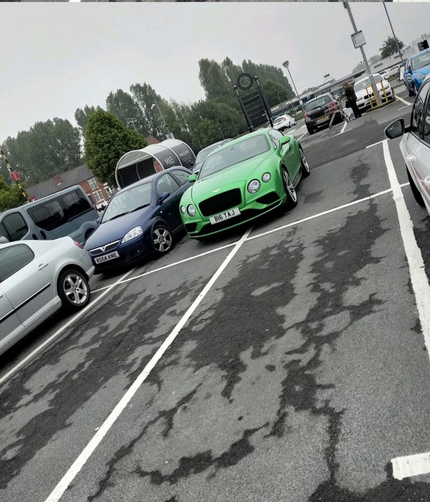 The BAD PARKING thread [vol4] - Page 518 - General Gassing - PistonHeads UK - The image depicts an outdoor parking lot with several cars. A distinctive feature of this scene is the presence of a bright green Bentley car parked in one of the spaces, which stands out due to its vibrant color and unique design. This luxury vehicle is surrounded by other cars, which appear to be of more modest makes and models, indicating a diverse group of vehicles in the lot. The ground appears wet, suggesting that it may have recently rained or that the area has been washed for cleaning purposes.