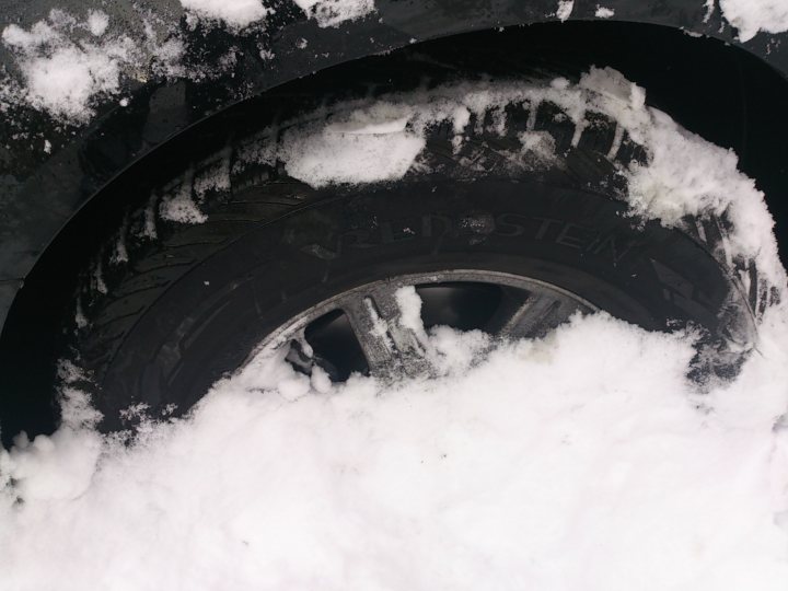 Winter tyres. - Page 1 - Off Road - PistonHeads - The image depicts a close-up view of a winter tire partially covered with snow. The tire bears a German-language brand name that is partially visible, with the letters "MOST" suggesting it's not entirely clear. The presence of snow mounds around the tire and on its sidewall indicates recent snowfall or ongoing snow removal efforts. The tire appears to be a standard type used on vehicles, and the winter tire's tread pattern is designed for traction on snowy surfaces.