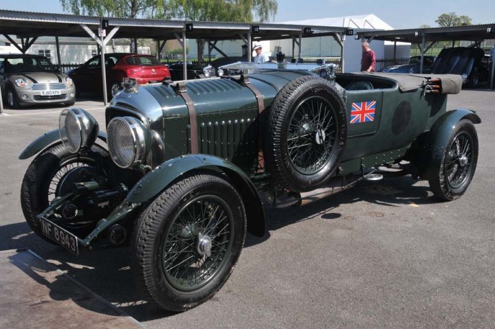 Goodwood yesterday...... - Page 1 - Classic Cars and Yesterday's Heroes - PistonHeads - The image displays an antique British racing car parked outside under sunlight. The car, with its distinctive round headlights and large spoked wheels, is painted a dark shade, possibly green or black. A noticeable emblem of a British flag on the side of the vehicle adds to its vintage charm. The car rests on solid wheels and appears to be in well-maintained condition. In the background, there are modern cars and a clear sky, suggesting the location might be a car show or a historical display setting.