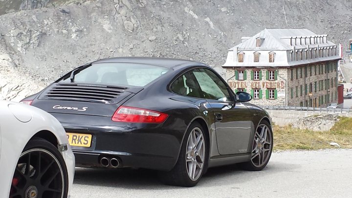 lowering a 997 - Page 3 - Porsche General - PistonHeads - The image shows a black Porsche sports car parked next to a mountainous area. The car is positioned on the left side of the photo, facing towards the right, and has the registration number "RK50 YK" visible on its license plate. In the background, there's another vehicle partially visible on the far left and some buildings that resemble small houses or possibly a small village or community. The terrain appears rocky with sparse vegetation, suggesting an alpine or mountainous region. There's no text present in the image.