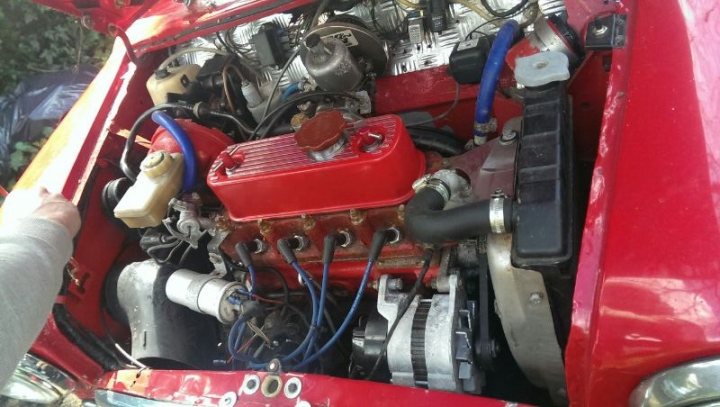 HELP please looking to engine swap or supercharge my engine  - Page 1 - Classic Minis - PistonHeads - The image showcases the engine of a red car. It's an engine that has seen better days, as it's been pitted against trees and rocks, indicating that it's been used off-road. The engine is dense with various mechanical components and parts. A clear plastic cover on the engine ensures that the mechanical components inside remain protected. There's also a person standing to the left of the image, possibly inspecting or working on the engine. The overall scene suggests an outdoor setting, possibly a garage or a shop, where the engine is being serviced or sorted.