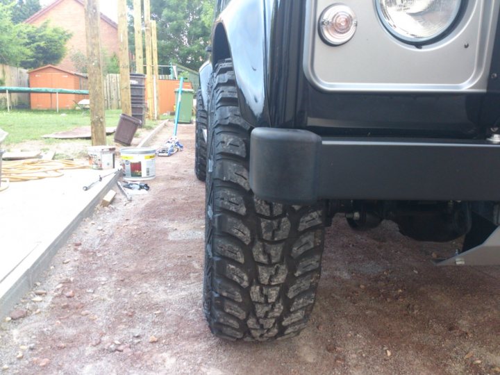 Wheel spacers on a Defender 110XS - Page 1 - Land Rover - PistonHeads - This image shows a gray off-road vehicle with large, knobby tires parked on a patchy dirt surface. A close-up view is centered on one front wheel, which is angled touching the ground. The vehicle appears to be a utility or work vehicle, possibly used for off-road or construction purposes. Behind the vehicle, there is a residential area with fences and a building. The foreground shows some materials, tools, and hand luggage, which are not in focus and appear unrelated to the vehicle.