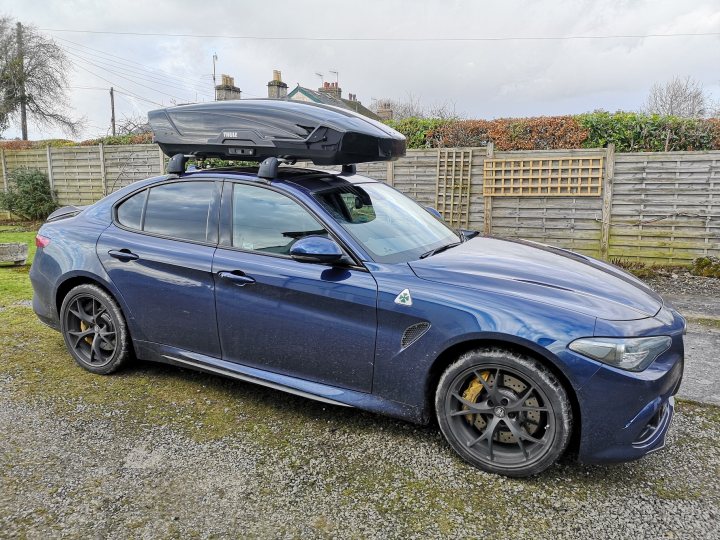 Alfa Romeo Roof Bars - Giulia QV - Page 1 - Alfa Romeo, Fiat & Lancia - PistonHeads - This image depicts a blue Alfa Romeo car parked outdoors, likely in a rural setting given the grass and open space. The car is topped with a large black luggage rack or roof rack. The vehicle's side mirrors are visible, and there's a noticeable sunroof as well. In the background, there's an indication of a fence or other boundary, suggesting this might be a private property or a parking area near homes. There is no text present in the image.