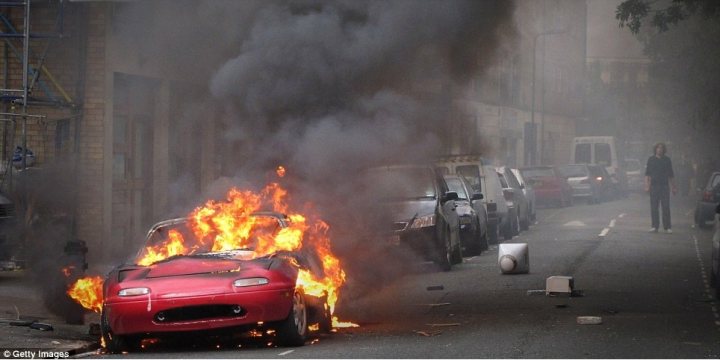 Badly modified cars thread - Page 57 - General Gassing - PistonHeads - In the image, there is a vibrant red sports car engulfed in flames on a city street. Smoke is billowing out of the hood and roof of the car, and the entire vehicle appears to be on fire. There are buildings in the background with visible signs of damage, indicating that the car's fire could have started there too. A lone pedestrian can be seen walking on the sidewalk, approaching the scene, possibly in shock at the unfolding event.