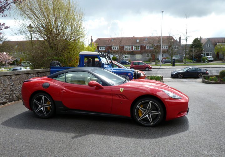 Scotland's Finest Spotted!! [Vol 2] - Page 85 - Scotland - PistonHeads - The image features a vibrant red Ferrari sports car parked on a paved surface. The car has a sleek design, with a prominent hood and side vents, characteristic of the brand's high-performance models. In the background, there is a blue utility pickup truck and a red hatchback car parked nearby. The setting appears to be a residential area, with houses visible in the distanced background. The sky is overcast with a diffused cloud cover, suggesting it might be a cool or cloudy day.