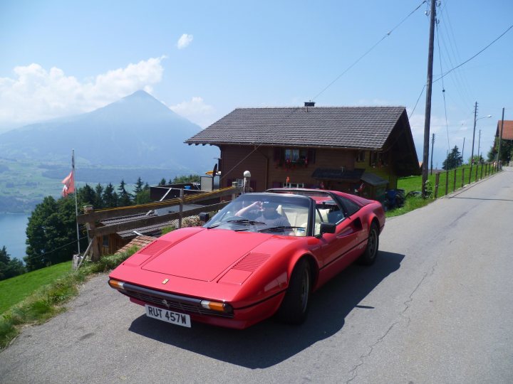 Pistonheads - The image shows a picturesque scene featuring a red sports car parked on a winding road. The car is sleek and has a convertible top up. It's parked next to a rustic wooden house with a shingled roof, surrounded by lush greenery and trees. The landscape suggests a mountainside location, and somewhere in the distance, beyond the car and house, there's a snowy peak and a small flagpole with a single flag flying on the left side of the image. Above this idyllic setting, the sky is clear with only a few scattered clouds. The scenery is serene and the setting appears to be in a mountainous area.