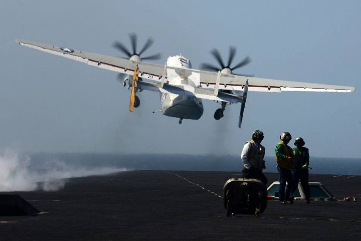 Post amazingly cool pictures of aircraft (Volume 2) - Page 203 - Boats, Planes & Trains - PistonHeads - The image shows a military cargo plane in flight, with its landing gear down. Below, on an aircraft carrier, there are personnel observing the aircraft's landing. The carrier appears to be spewing steam, possibly due to the heat generated by the approaching plane. The sky above the carrier is clear, and there is a person wearing protective eyewear on the deck, likely to shield from the jet blast. The scene conveys military personnel performing a task or exercise.