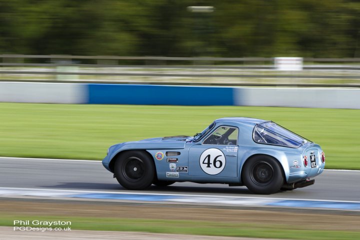 Early TVR Pictures - Page 55 - Classics - PistonHeads - The image showcases a vibrant blue racing car on a curvy track. The car is seen in action, tilting to the right as it navigates the bend. The setting appears to be a sunny day, with clear visibility and a professional racing environment. There is also a photographer's credit at the top of the image, indicating that this is a photograph taken for design purposes.