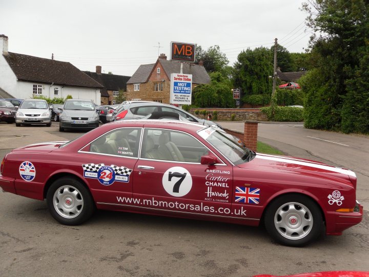 Stickered Pistonheads - This image features a vintage red car with a distinctive design, parked in front of a white house with green accents. The car is partially obscured by the visibility of both the front and the back due to the perspective from which the photo is taken. The vehicle is adorned with a checkered pattern and the number 7, as well as text that reads "Charles Chell" and "The Great British Breakfast." There are stickers present, including "24" and "www.mbmmotorsales.co.uk." The background reveals a residential area with parked cars and trees, suggesting a peaceful suburban setting.