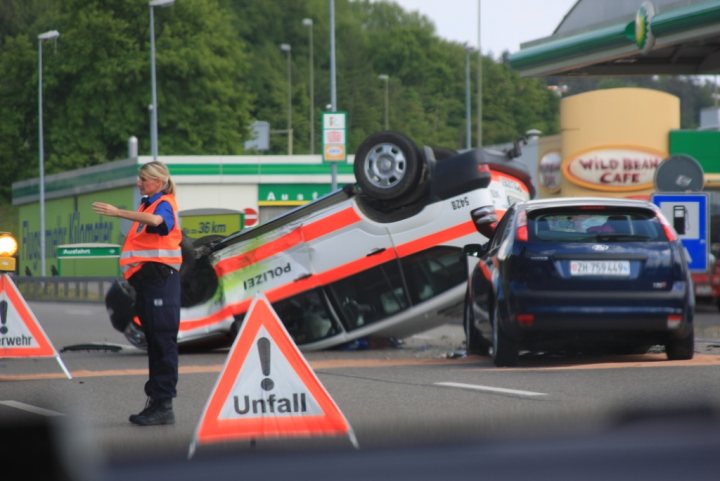 Airport Pistonheads Zurich Whoops Police - The image captures a crash scene on a highway. A red, white, and orange car has flipped over on its side, and the driver is reported dead at the scene. A police officer is gesturing to nearby motorists, trying to ensure their safety. The motorists are passing the crash site, which is marked by a blue sign with an orange exclamation symbol. The environment suggests it's either late in the day or early morning, given the muted lighting conditions.