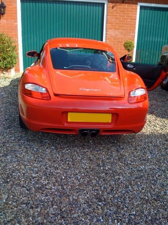 Pistonheads - This image showcases a distinctive orange sports car parked in front of a brick building, which has green wooden garage doors. The car is positioned facing the viewer, with the license plate prominently visible, suggesting it's a well-maintained vehicle. The background reveals a typical suburban scene, with another car parked nearby and a tree adding a touch of nature to the setting. The contrast between the aerodynamic design of the sports car and the traditional aesthetic of the garage doors creates an interesting juxtaposition.