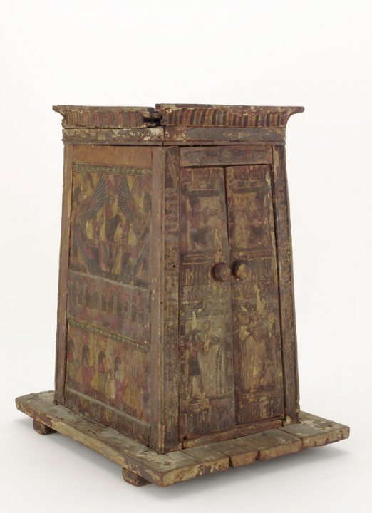 This image shows a vintage furniture piece, specifically what appears to be an old, possibly antique wardrobe or cupboard. The structure has a rich, dark finish that seems to be ageing, and it is decorated in brown and beige tones that suggest a patina of time. Each panel of the wardrobe features intricate patterns and possibly depicts figures or scenes. The cupboard has visible damage and signs of wear, which adds to its rustic and historically significant appearance. The wardrobe remains stationary, with its stand visible. The background is neutral and does not detract from the piece itself.