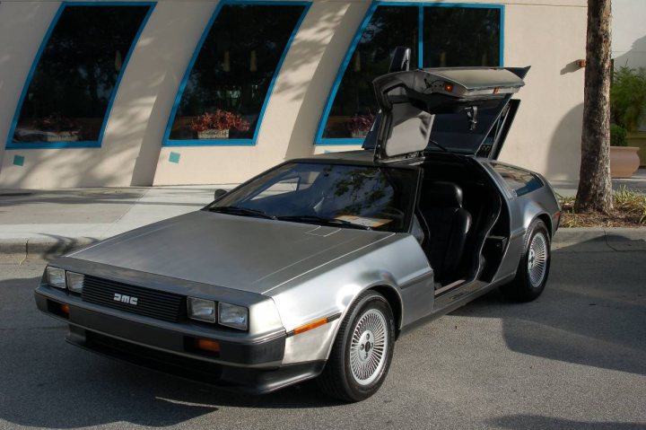 Delorean Piston Heads Pistonheads - This image captures a silver metal-edged DeLorean sports car parked outside a building with multiple rounded windows. The car's doors are open, suggesting it is currently not in use. The vehicle is on the driver's side of the street, indicating a right-hand drive side. The background shows the facade of the building and some vegetation, adding context to the scene. There are no people visible in the image.