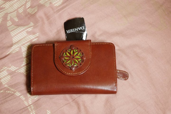 The image features a brown wallet with a visible imprint of a burst light motif on its surface and a key chain attached to it towards the top. It is positioned on a fabric, which has a pink hue and a white pattern, possibly a bedsheet or blanket. The wallet has tanned leather straps at the top for carrying or attaching to other belongings. The focus of the photograph is on the wallet, and the background is out of focus.