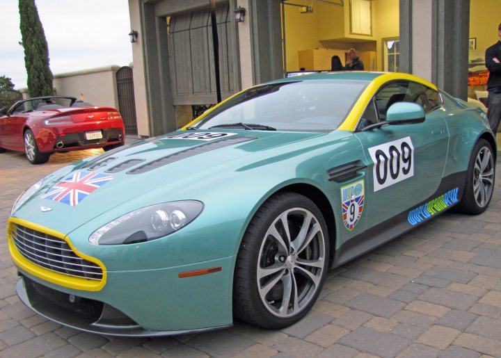 Pics from Aston event, Monterey - Page 1 - Aston Martin - PistonHeads - The image features a distinctive blue Aston Martin sports car parked on a brick driveway. The car is adorned with a striking yellow stripe and various British flags, suggesting a theme of national pride. The number 09 prominently displayed on the side, adding a personalized touch to the vehicle. In the background, several other cars and individuals can be seen, indicating a social gathering or event of some sort. The setting appears to be at a show car pavilion, where the Aston Martin stands out as the main attraction.