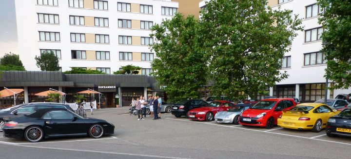 Geneva road trip & tour of JLC Manufacture  - Page 10 - Aston Martin - PistonHeads - The image shows a well-maintained, urban parking lot. There are several cars parked, including a variety of red, black, yellow, and blue models, which are neatly lined up along the white parking lines. The presence of signs and access points indicates a regulated parking area. The environment suggests an area outside a large building or complex, as evidenced by the white buildings in the background. The scene is likely captured during the day, given the clear visibility and lighting conditions.