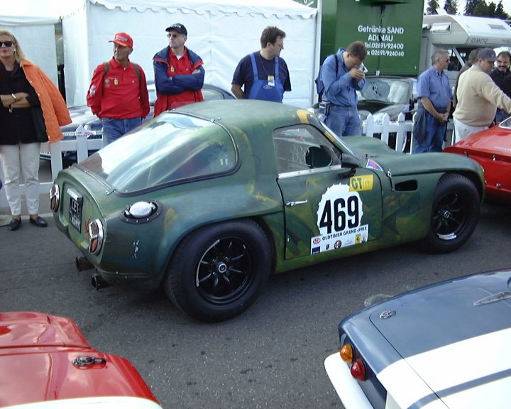 Early TVR Pictures - Page 19 - Classics - PistonHeads - The image captures a vintage car show scene. A restored green 469 race car is the central focus, occupying a significant portion of the image. The car is adorned with the number "469", and the door displays a logo, possibly signifying the brand or team using the vehicle in the past. Various onlookers are present in the image, observing the show car and several other cars on the adjacent road. They are dressed casually, with some carrying backpacks and handbags. The atmosphere suggests a casual day out for car enthusiasts.