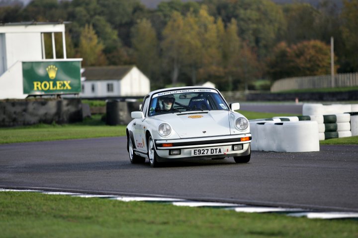 Octane magazine track day 3rd.Nov.  - Page 1 - Goodwood Events - PistonHeads - The image is a photograph featuring a vintage or classic car driving on a racetrack. The vehicle appears to be a Porsche 911 GT2 RS given its distinct design, evidenced by the long hood, short rear deck, and large rear wing. The car is labeled with the registration number "E52 DTA". The track is bordered by a series of hay bales, and stands lined with seating and a promotional advertisement can be seen in the background. The image conveys an atmosphere of motorsport and racing activity.