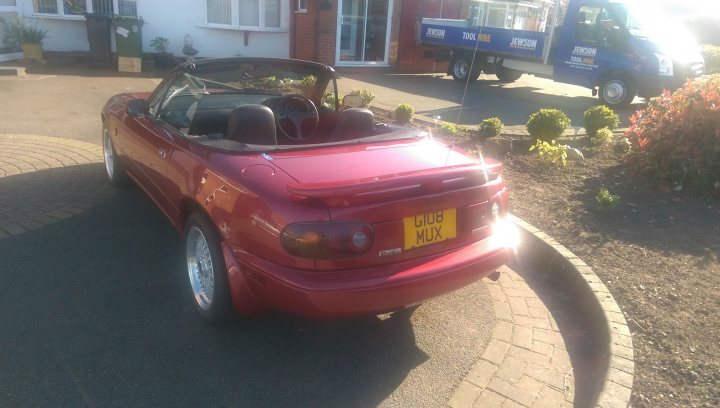 My new Eunos! - Page 1 - Mazda MX5/Eunos/Miata - PistonHeads - The image depicts a vibrant red convertible sports car parked on a driveway. The car appears to be in good condition, with a freshly applied coat of paint. It's a bright sunny day, with the sun shining directly on the car, accentuating its color and creating a picturesque scene. In the background, there are other vehicles, including a red truck, suggesting this might be a residential area with various types of vehicles parked. Despite the various vehicles, the convertible sports car is the prominent object in the image.