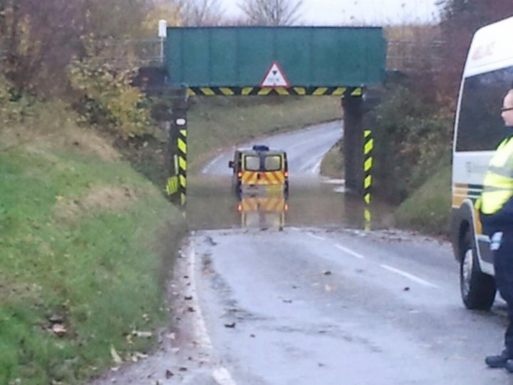 Doh!! - Page 1 - Northamptonshire - PistonHeads - In this image, a construction site area can be seen with water flooding the road. A large green bridge that spans the height of the road crosses over a hurricane railing. A yellow car with a police light on the roof sits in the midst of the flooded road. The road itself is low, and the bridge seems to be a route for construction or possibly evacuation. In the far right corner, a person wearing a bright yellow vest stands, possibly observing or supervising the situation.