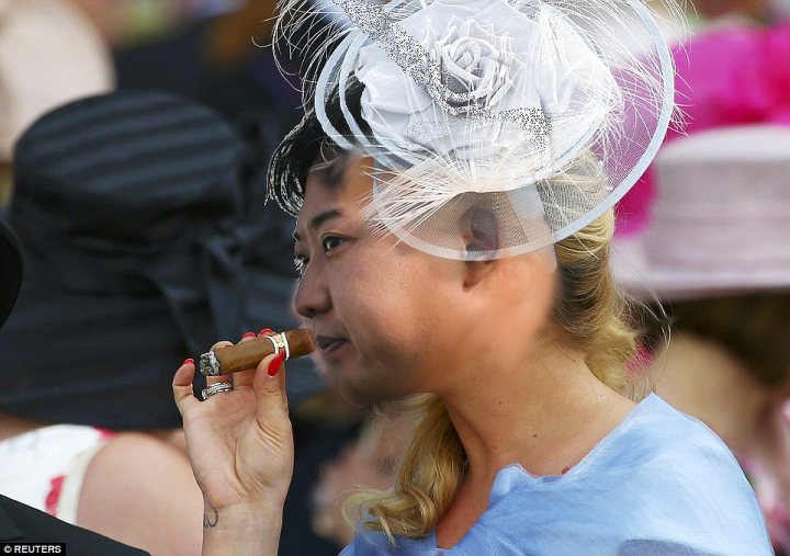 North Korea photoshop contest - Page 29 - The Lounge - PistonHeads - In the image, a woman is the central figure, poised elegantly against a blurred background. She is adorned in a wide-brimmed hat, complete with a large bow and white feathers on the side. The hat's white color contrasts beautifully with her outfit, adding a touch of sophistication.

In her right hand, she holds a cigar, bringing it to her mouth. She appears to be in the midst of taking a puff, adding a layer of mystery to her persona. 

Her body faces the camera but her head is turned towards the left, suggesting that she might be looking at someone or something out of view. Her overall demeanor exudes a sense of calm confidence and charm. 

The background, though blurred, seems to be filled with other people, possibly in a social setting. However, due to the focus on the woman, specific details about them are not clearly discernible. The woman's stylish attire and behavior suggest a stylish and well-put-together event.