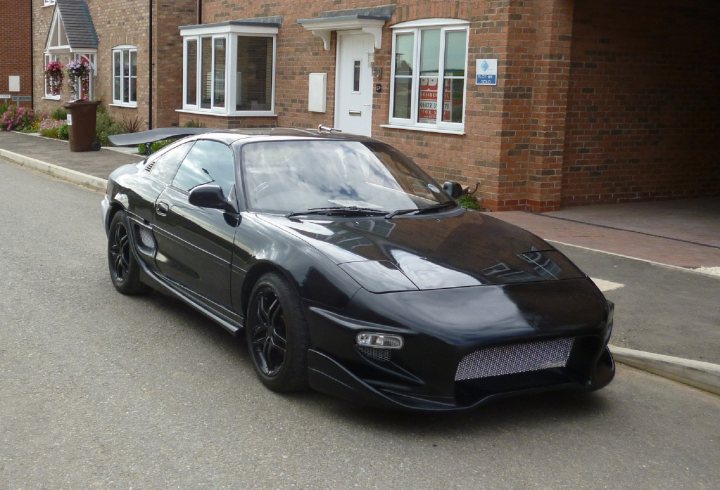 MR2 owners - How many have you owned? - Page 4 - Jap Chat - PistonHeads - The image shows a luxurious black sports car parked on a concrete driveway in front of what appears to be a residential house. The car exhibits a sleek, aerodynamic shape with smooth, glossy surfaces. An open-style front grille stands out, and there are visible elements of air intakes and possibly wheels, suggesting high performance capabilities. Interestingly, there is also an emblem on the front bumper that resembles a lightning bolt or some form of speed symbol. It seems to be a calm, suburban environment, with no other vehicles or people immediately visible in the image.