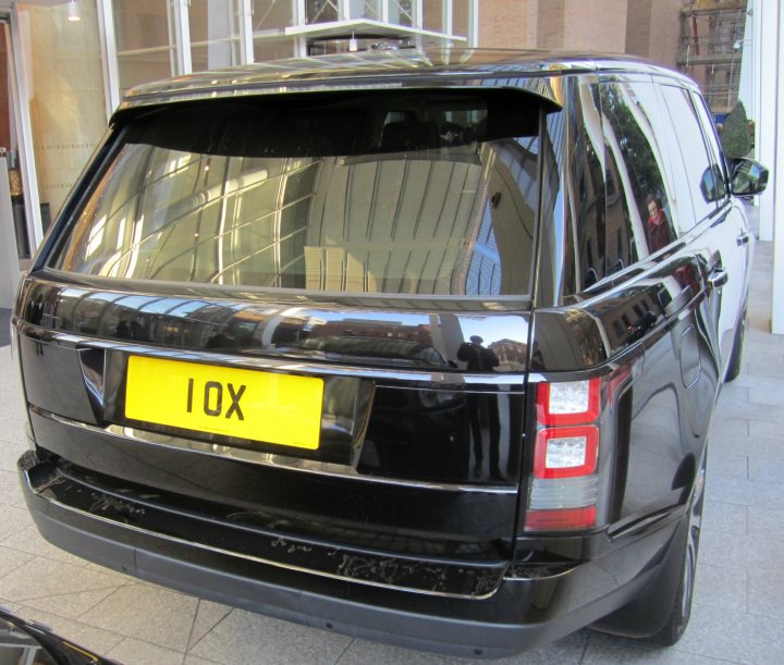 Real Good Number Plates : Vol 4 - Page 274 - General Gassing - PistonHeads - The image shows the rear view of a black SUV parked outdoors. The vehicle is sleek and modern in appearance, with a glossy finish on the black paint. The SUV displays a yellow license plate with the letters "Iox" prominently displayed in the center, which suggests it could be a taxi or ride-sharing vehicle due to the unique license plate configuration. The design of the rear lights is modern and rectangular, signaling the car's up-to-date styling. There is a reflection of trees on the car's window, indicating that the car is parked near a wooded area. The backdrop to this scene appears to be a row of buildings, suggesting the vehicle may be parked on the side of a road.