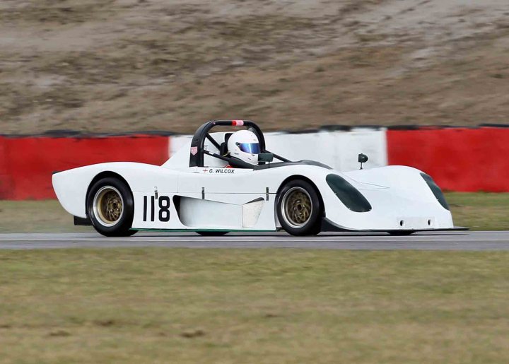 Buy Pistonheads - The image captures a dynamic race track scene featuring a sleek, white racing car. The car, labeled with the number "118" and the name "G. Willcock," is positioned on the left side of the frame. It's moving on a racing track with green grass in the foreground. The track's background is dominated by white barriers, providing a sense of depth and motion.