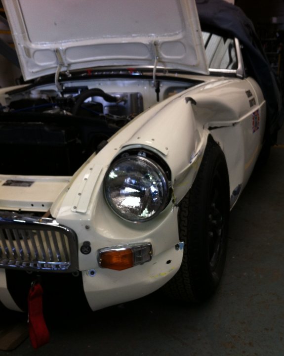 Early MGB front wing - Page 1 - MG - PistonHeads - The image shows a classic, vintage car with its hood open. The car appears to be an older model, potentially designed in the late 20th century, and its hood is propped up with a prop. The vehicle's hood is directly above the car's engine bay, displaying standard components including an alternator, battery, and various hoses and wires. The car's grill features headlights and a distinctive emblem, indicative of its brand. The bumpers seem to be damaged, and there are no visible brands or distinctive features that would suggest a specific make or model. The car is parked indoors, with a green floor visible beneath it.