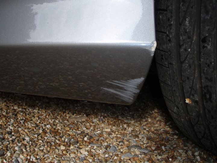 Front bumper respray - Page 1 - Aston Martin - PistonHeads - The image captures a close-up view of a vehicle, focusing on the front portion of its fender. The fender shows signs of wear and tear, most notably a significant scratch marking its surface. The vehicle is parked on a gravel surface, as evidenced by the scattered stones that form the backdrop of the image. The tire of the vehicle is also visible, suggesting that the car is stationary at the time the photo was taken. The image provides a clear view of the vehicle's condition and the environment it's parked in.