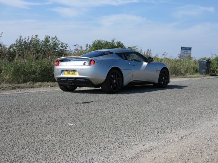 I've also bought one.... - Page 1 - Evora - PistonHeads - The image shows a sleek, silver sports car parked on the concrete side of a road. The car is positioned in front of a green sign with a white border that has some text on it, although the resolution does not allow for clear legibility of the text. To the right of the sign, there is a gray trash bin. The background features a sky filled with thin clouds, suggesting it might be a cloudy day. The car shines under the light, indicating a well-maintained vehicle.