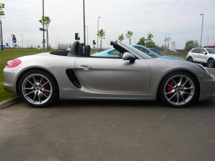 981 Boxster S Spec - Thoughts? - Page 6 - Porsche General - PistonHeads - The image shows a light gray Porsche sports car parked on an asphalt surface. The car has a black convertible top closed and features the iconic Porsche badge on the front hood. It's parked at an angle, with the rear wheel towards the left of the frame and the front wheel angled towards the right. The background is relatively nondescript with street lamps and other parked cars visible, suggesting an urban or suburban setting.