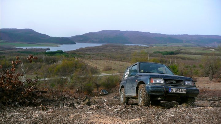 background car pics :) - Page 15 - Readers' Cars - PistonHeads - The image depicts a scene of three distinct landscapes combining into one. In the foreground, a dark-colored SUV is on a dirt road, slightly angled towards a hill, suggesting it might be climbing. In the middle ground, a large body of water stands out, its calm surface reflecting the distant mountains. Beyond the water, a rugged mountainous terrain rises, marking the distant horizon. The overcast sky suggests a cool or possibly unsettled climate.