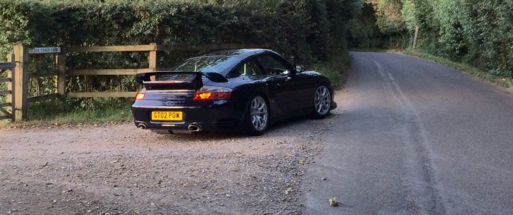 GT2 - Page 111 - Porsche General - PistonHeads - The image shows a black sports car parked on the side of a country road. The car's rear end is clearly visible, displaying a large rear spoiler and the characteristic lines of a sports car design. The surrounding environment is rural, with a fence and lush greenery visible. The sky is not visible in the frame, but the overall setting suggests a calm, isolated location. There is a yellow license plate on the car.