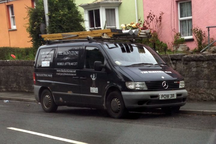 Real Good Number Plates : Vol 4 - Page 1 - General Gassing - PistonHeads - The image depicts a black van parked on a street by the side of a building. The van is equipped with a ladder on top, with a number of pipes or cylinders attached to it. On the side of the van, there is printed text and a logo, indicating it might be a work vehicle, possibly for a business or organization. To the left of the photo, there's a glimpse of a vibrant yellow building, adding a pop of color to the scene.