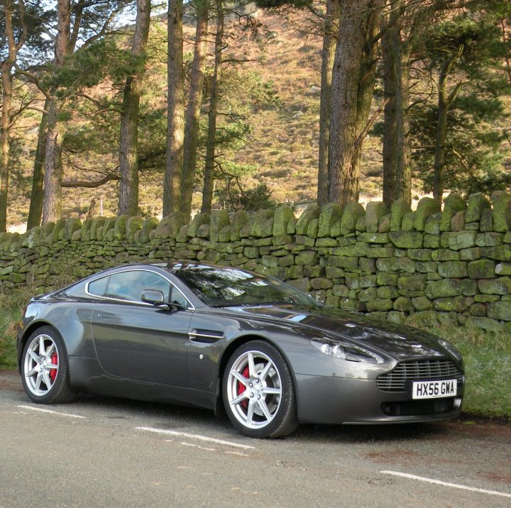 V8 Vantage - 3 week / 700 mile update - Page 1 - Aston Martin - PistonHeads - The image shows a black Aston Martin car parked at the roadside next to a natural stone wall. The car features red accents on its alloy wheels. In the background, there is a lush green hillside with mature trees that seem to provide a secluded environment. The sky is visible, suggesting it's a sunny day. The overall setting gives the impression of a calm and peaceful location.