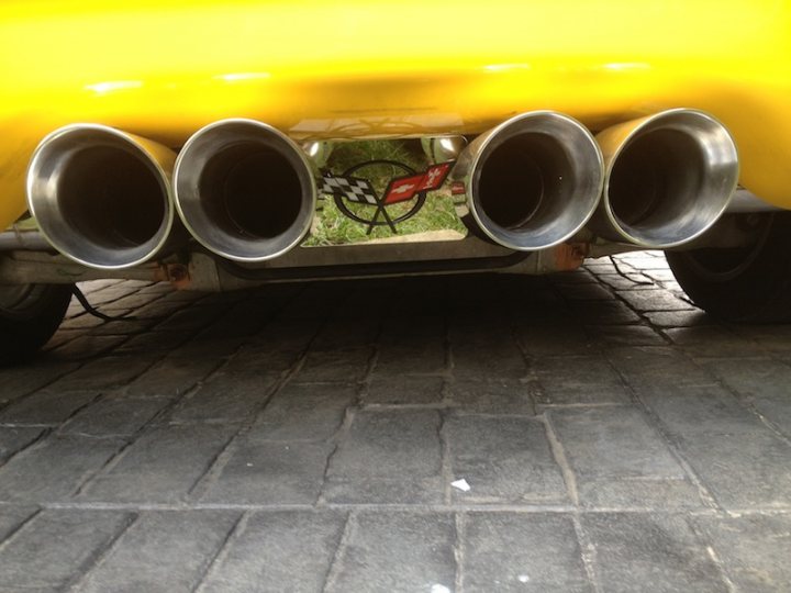 A blue and yellow fire hydrant sitting on the side of a road - Pistonheads - The image features a close-up view of a yellow car's rear bumper. Two dual tailpipes are prominently visible, with one clearly showing a two-leaf clover logo. The car's back bumper also displays a racing flag graphic, alternating red and green. The photograph is taken from an elevated angle, allowing the viewer to see under the vehicle. The setting appears to be on an outdoor cobblestone surface, as indicated by the open and gridded texture of the ground.