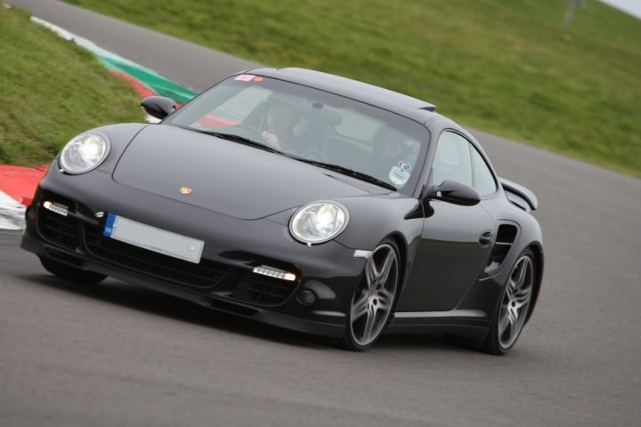 Pictures of 997 turbo's - Page 1 - Porsche General - PistonHeads - The image depicts a black sports car in motion on what appears to be a race track. The vehicle is sleek and designed with aerodynamics in mind, possibly for performance. The car's headlights are on, suggesting it's either during the daytime under overcast conditions or in the evening. The road conditions show signs of wear, with some rubber marks, indicating frequent use or perhaps racing tires. The car's license plate is partially visible at the front, though the text is not legible.