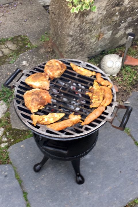 Photo of your dinner (vol 2) - Page 283 - Food, Drink & Restaurants - PistonHeads - The image shows a grill filled with various pieces of chicken. A medley of chicken legs, wings, and possibly breasts are arranged on the grill, which is set atop a stone patio. The grill appears to be of a charcoal type, spread with ashes, indicating it has been used. In the background, there are green plants and a rock wall with raised mounds, adding a natural element to the outdoor setting. Surrounding the grill is a cluttered area with items on the ground, suggesting a casual outdoor cooking environment.