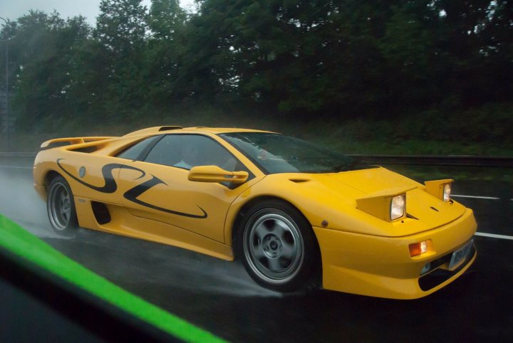 V12 Lambo Photo Thread - Page 3 - Diablo/Murcielago/Aventador - PistonHeads - The image features a yellow sports car on a road, presumably in motion. The car is sleek and low, with a distinctive paint scheme and a large rear wing. The weather conditions seem to be rainy, as evidenced by the misty raindrops on the windshield and the wet road surface. The background is slightly blurred, emphasizing the speed and dynamism of the vehicle. A green stripe on the side of the car adds a touch of contrast to its design.