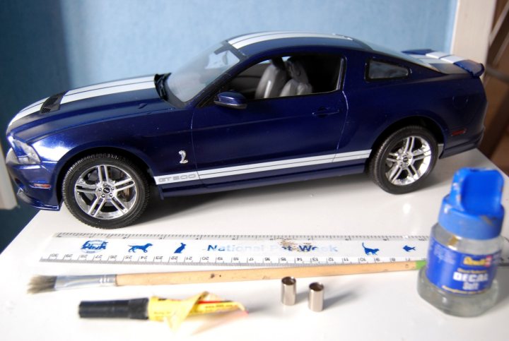 Revell 1/12 Ford Shelby GT500 - Page 3 - Scale Models - PistonHeads - The image features a vibrant blue Mustang model car, meticulously assembled on top of a white ruler. The toy car, which is detailed with the iconic stripes and the Ford logo, is a testament to careful craftsmanship, as evidenced by its pristine condition. A few tools, including scissors and glue, are strategically placed next to it on the table, suggesting the process of building or modifying this model car. The overall scene suggests a hobbyist's workspace, filled with precision instruments and carefully chosen models.